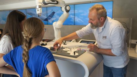 Alexander Kleic tourt mit dem M+E Infotruck durch Bayern. Hier erklärt er zwei Schülerinnen, wie der Roboter funktioniert.
